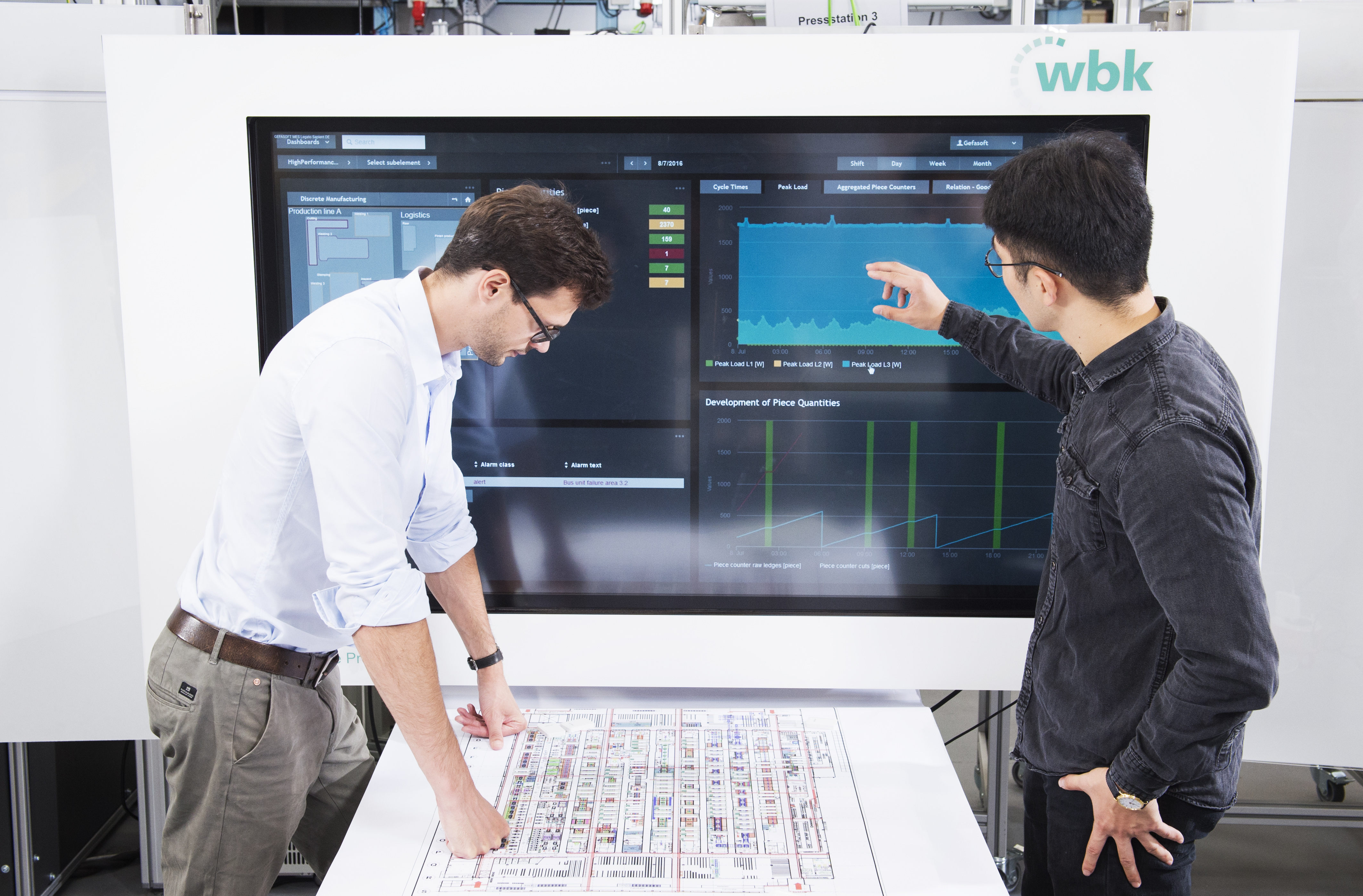 Zwei Fachkräfte analysieren Produktionsdaten auf einem großen Touchscreen in der Lernfabrik des wbk am KIT.