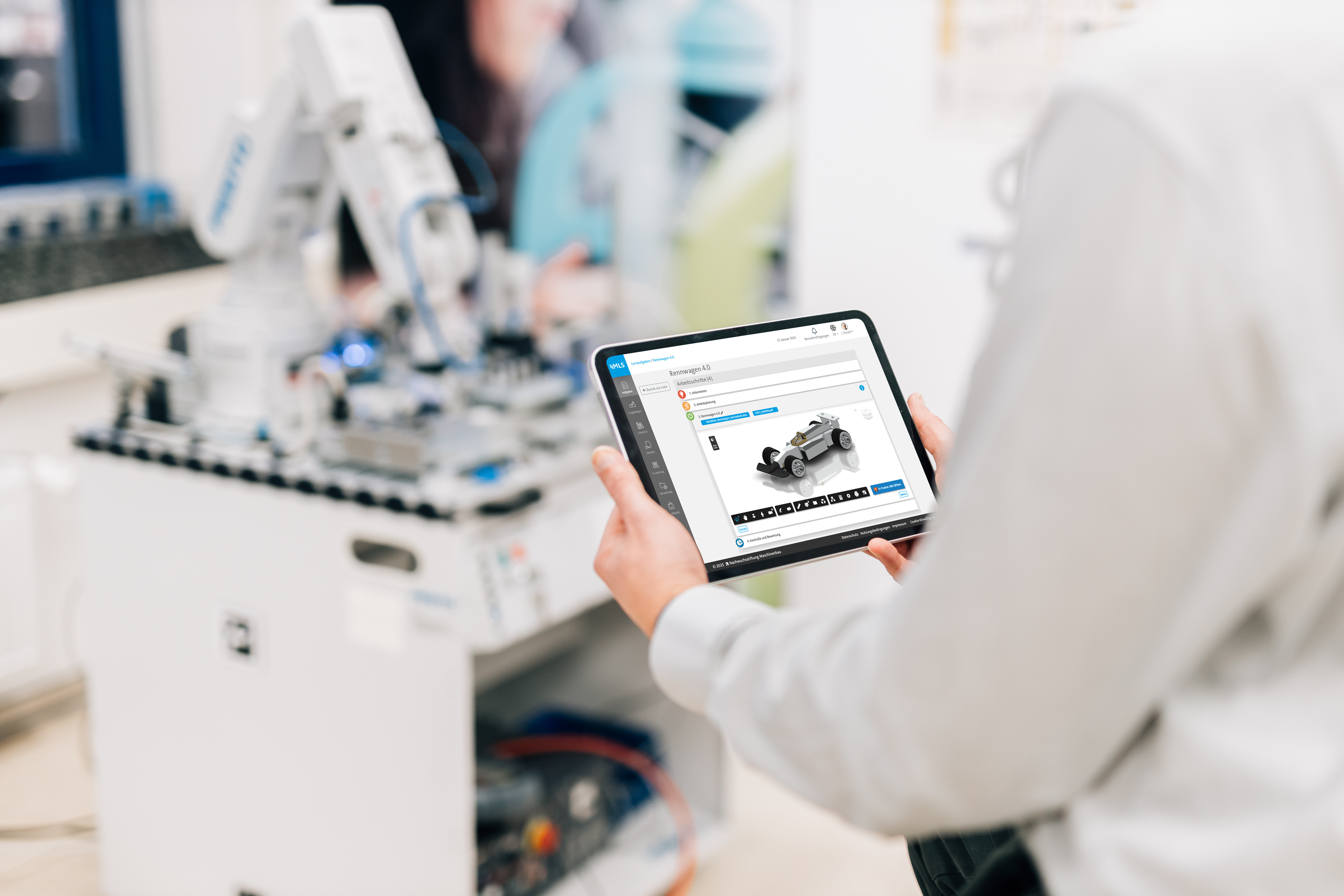 Person hält ein Tablet mit geöffneter Lernplattform MLS; auf dem Bildschirm ist eine Lernaufgabe zur Baugruppe Rennwagen sichtbar. Im Hintergrund eine technische Lernstation mit Roboterarm.