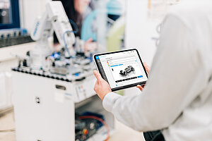 Person hält ein Tablet mit geöffneter Lernplattform MLS; auf dem Bildschirm ist eine Lernaufgabe zur Baugruppe Rennwagen sichtbar. Im Hintergrund eine technische Lernstation mit Roboterarm.