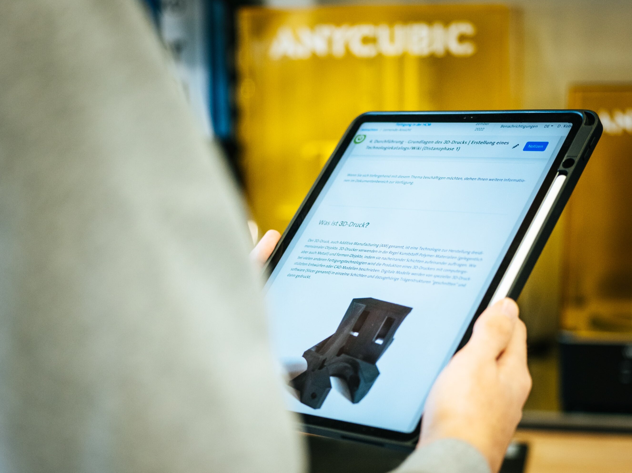 Person hält ein Tablet. Im Display ist die Lernplattform MLS mit einer Lernaufgabe zum Thema 3D-Druck zu sehen. Im Hintergrund eine Anycubic 3D-Drucker-Werkstatt.